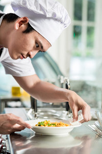 Young male culinary professional in a white chef's hat putting the finishing touches on a plated dish of food in a white bowl