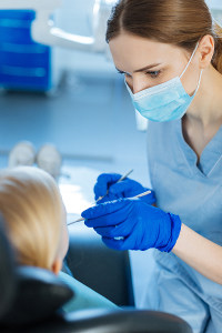 Dental assistant in blue scrubs and mask examining a young patient's teeth in a modern dental office
