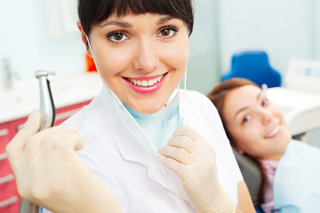 Dental hygienist in white coat with dental tools in hand, patient in background in dental clinic.