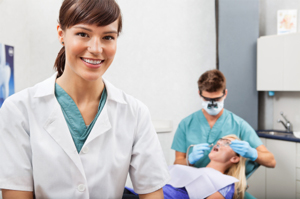 Dental hygienist in lab coat standing in dental office with dentist treating patient in background.