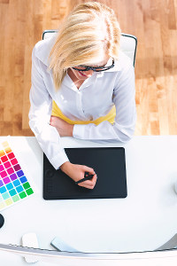 Professional graphic designer using a digital tablet with a stylus and color swatches on desk in a bright office setting