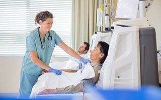 Female dialysis technician putting a blanket on a male patient who is sitting next to dialysis machines and another patient