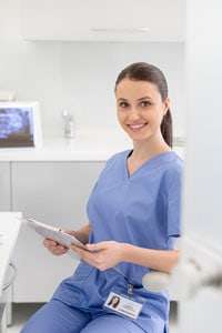 Young woman in blue scrubs smiling and holding a clipboard while sitting in a room of a medical facility