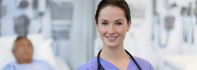 Smiling young woman wearing scrubs and a stethoscope around her neck standing in front of a patient in a hospital bed