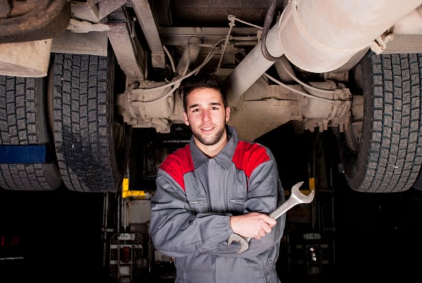 Diesel mechanic in gray and red uniform holding a wrench, smiling, standing under a lifted truck in a workshop.