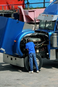 Diesel mechanic in blue uniform inspecting engine of a blue semi truck with open hood in a parking lot.