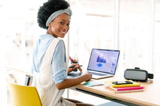 Young woman in casual attire working at a bright office desk with a laptop showing graphs, smiling at the camera, with digital gadgets and stationery around.