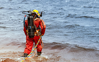 Rescue diver in orange suit and yellow helmet preparing to enter the water from the shore with ropes attached