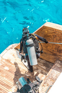 Scuba diver wearing full gear including a tank and fins preparing to enter the water from the back of a wooden boat