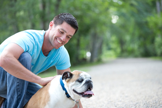 Smiling man in jeans and a blue T-shirt kneeling down and petting a Bulldog on an outdoor walking path