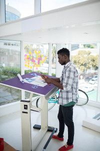 Young man using tablet at standing desk in a bright office space overlooking bicycles outside through large windows.