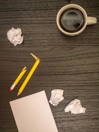 Top-down view of a workspace with crumpled paper balls, broken pencils, a cup of coffee, and a blank notepad on a dark wooden surface.