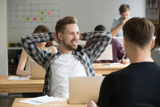 Relaxed young male professional with hands behind head smiling at colleague in busy office environment.