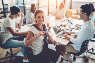 Young professionals collaborating in a brightly lit office, with a woman smiling at the camera and her colleagues working on laptops in the background.