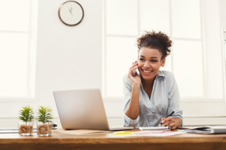Woman using laptop and talking on phone in a bright home office with paperwork, plants, and a wall clock.