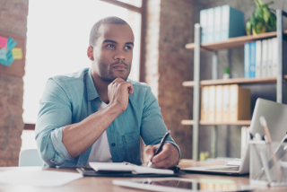 African American man sitting at desk in office, pensively looking away with chin resting on hand, laptop and notepad in front of him