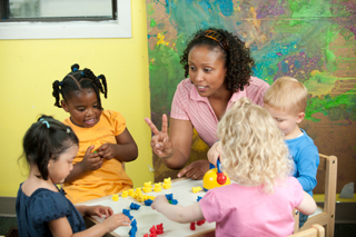 Diverse group of preschool children engaged in play at a colorful table with a female teacher guiding and interacting with them in a classroom setting.