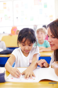 Young female teacher assisting a little girl with her workbook in a colorful elementary school classroom, with other students in the background.