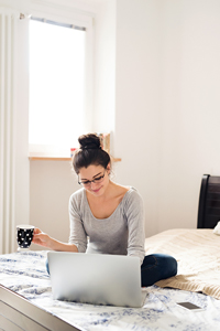 Woman in a gray top sitting on bed with laptop and holding a coffee mug while looking at screen