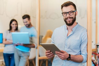 Man with glasses holding a tablet in an office environment with two colleagues working on laptops in the background.