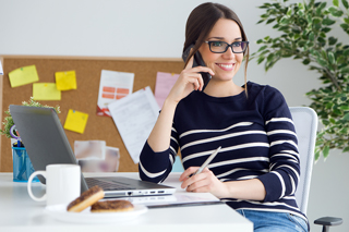 Smiling woman talking on the phone while working on her laptop in a home office setup.