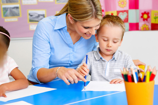 Teacher assisting young boy with writing at classroom desk, with colorful pencils on the side.