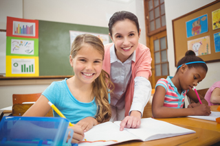 Teacher assisting students in elementary school classroom, with a focus on a smiling girl and teacher looking at the camera while another student works on her assignment.