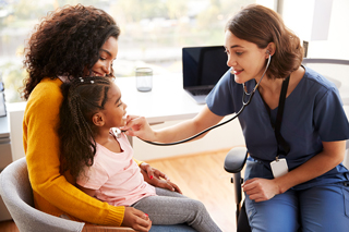 EKG technician using a stethoscope on a young girl sitting on her mother's lap during a medical consultation in a clinic, with natural light coming through large windows.