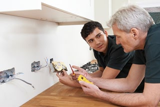 Two electricians, a young man and an older man, working together on wiring inside a kitchen, using a multimeter to test an electrical outlet.