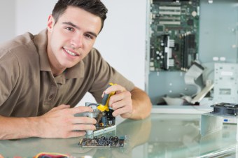 Smiling young man sitting at a worktable and using a torque screwdriver on a piece of computer hardware