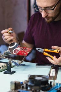 Man with glasses soldering a circuit board on a workbench using a magnifying glass and soldering iron.