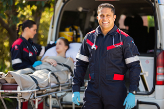 Paramedic smiling at camera outside ambulance while colleagues assist patient on stretcher in background.