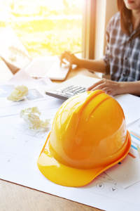 Person working at a desk with architectural plans and a laptop, with a focus on a yellow hard hat in the foreground.