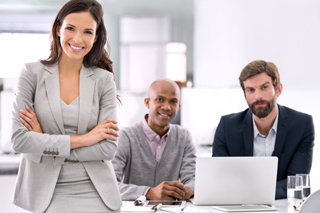 Three professional colleagues in a meeting, with a woman in a gray suit standing arms crossed smiling, flanked by two seated men, one looking at a laptop, in a bright office setting.