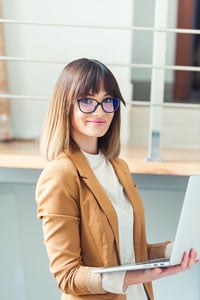 Professional woman in glasses holding a laptop in an office setting, smiling at the camera.