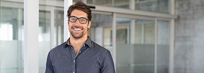 Smiling man wearing glasses and a dark-gray collared shirt standing in front of an office space with lots of windows