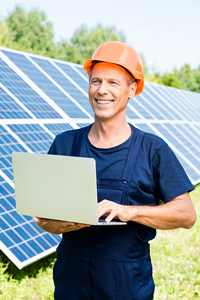 Technician in overalls and hard hat using laptop in front of solar panels