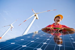 Engineer in safety vest and helmet inspecting solar panel with wind turbines in background under clear blue sky