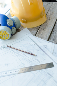 Construction blueprint plans with pencil, ruler, hard hat, and safety glasses on a wooden surface