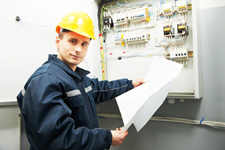 Male electrician in navy coat and yellow hardhat standing at electrical panel holding papers