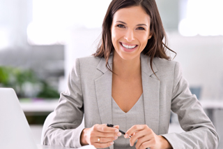 Businesswoman in gray blazer sitting at desk in office with laptop and pen in hands.