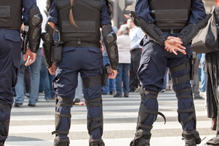 Police officers in riot gear standing in a line during a public event, equipped with body armor and tactical equipment.