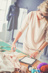Fashion designer measuring fabric with a tape measure at a workshop table, using a digital tablet, surrounded by sewing accessories.