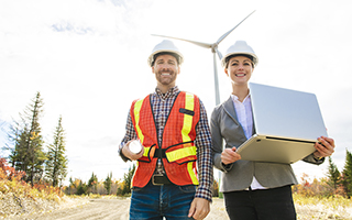 Two engineers in safety gear with a laptop and hard hats standing in front of a wind turbine in a rural landscape