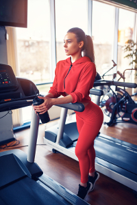 Young woman in red sportswear holding water bottle while standing on treadmill in gym