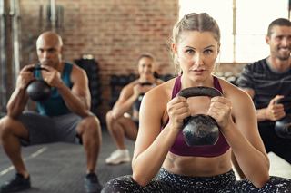Group fitness class with diverse adults using kettlebells in a brick-walled gym, focusing on woman in front holding a kettlebell with determination.