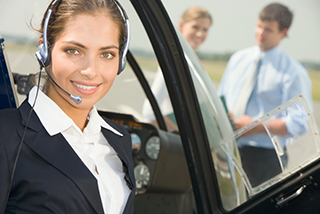 Female pilot with headset smiling in helicopter cockpit, with two people talking in the background on the tarmac.