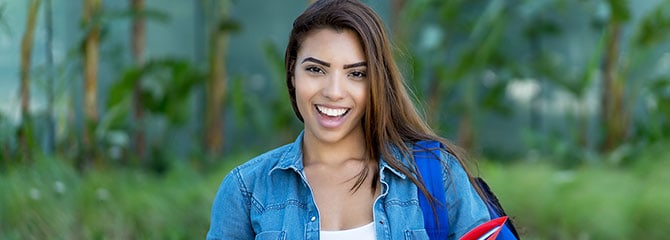 Young woman in a jean jacket smiling, carrying a red folder, and wearing a backpack on one shoulder while standing outside