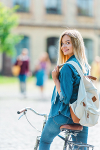 Young woman with blonde hair sitting on a bicycle, wearing a denim jacket and carrying a backpack, looking over her shoulder with a smile in an urban setting.