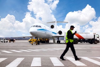 Airport worker walking across the tarmac with airplane, fuel truck, and service vehicles in the background under a cloudy sky.